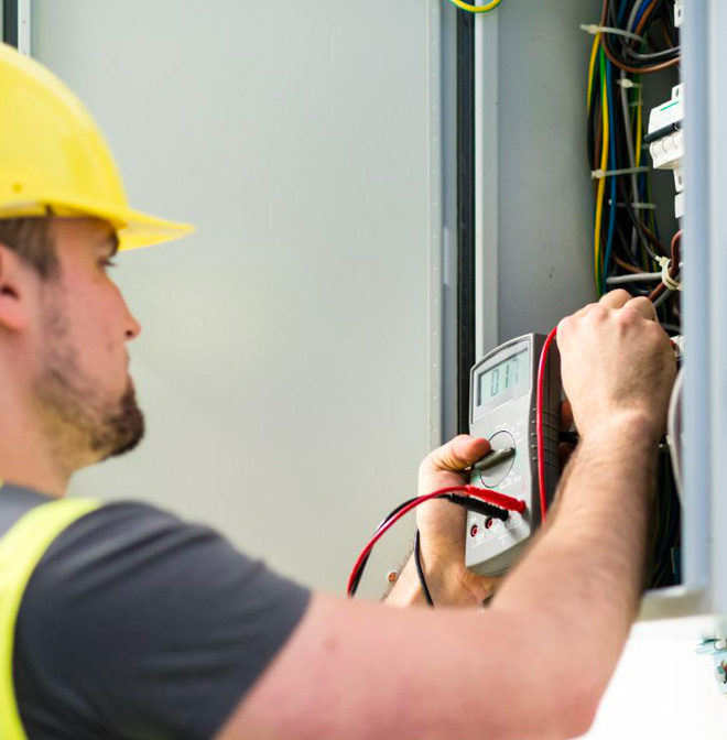 Un électricien, portant un casque jaune, mesure la tension dans un tableau électrique